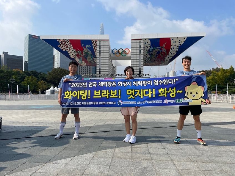 올림픽공원 평화의 광장 앞에서 화성시 체력왕 대표로 전국대회에 참여한 시민 3명이 현수막 을 사진을 찍고 있다. (화성도시공사 제공) ⓒ타임즈